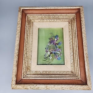 May include: Framed floral artwork featuring a bouquet of purple, blue, and white flowers on a green background. The artwork is set within a multi-layered frame with brown and cream tones, creating a vintage aesthetic.
