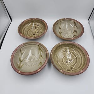 May include: Four ceramic bowls with a light brown interior and a dark brown rim. Each bowl features a unique design with white and brown accents, including spiral and floral patterns. The bowls are arranged on a white surface.