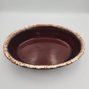 Pode incluir: Uma assadeira oval de cerâmica com um interior profundo de cor castanho-escuro brilhante. A borda larga e elevada apresenta um padrão decorativo de esmalte texturizado em tons de creme e castanho-claro, criando contraste com o centro.