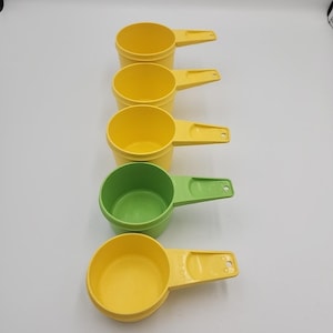 Può includere: Un set di cinque misurini colorati. Quattro sono gialli e uno è verde. Ogni tazza ha un manico. Le tazze sono di plastica e sono disposte in fila, con la tazza verde al centro.