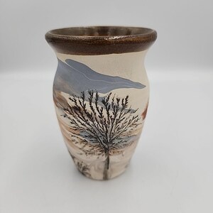 May include: A ceramic vase with a landscape design. The vase features a cream-colored base with a brown rim. A detailed illustration of a tree is painted on the front, with a blue and brown landscape in the background.