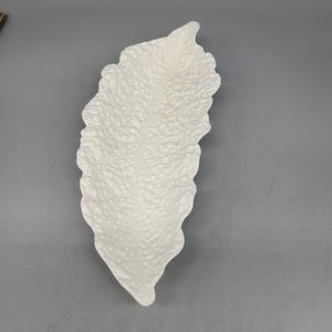 May include: A white, leaf-shaped decorative dish with a textured surface. The dish has a long, slender shape with a slightly raised edge. The surface has a pattern of small, irregular bumps. The dish is made of a solid, opaque material.