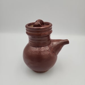 May include: A small, round, dark brown ceramic vessel with a textured surface, a short spout on the side, and a matching lid featuring a small knob handle. The piece has a glossy finish and a distinct neck section with horizontal ridges.