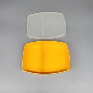 May include: A yellow rectangular food container with a clear lid. The container is divided into two sections. The lid is clear and has a circular indentation in the center. The container is on a gray surface.