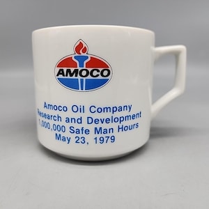 Puede incluir: Taza de cerámica blanca con el logotipo de Amoco en rojo, blanco y azul. La taza incluye el texto: "Amoco Oil Company Research and Development 1,000,000 Safe Man Hours May 23, 1979". La taza tiene un asa cuadrada.