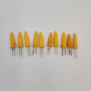 Puede incluir: Una colección de diez sujetadores de mazorcas de maíz amarillos con púas de metal plateado. Los sujetadores de maíz tienen forma de mazorcas de maíz en miniatura y están dispuestos en fila sobre un fondo blanco. Probablemente se utilizan para servir mazorcas de maíz.