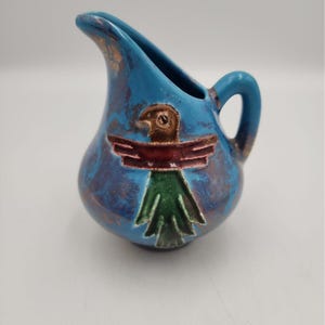 May include: A small ceramic pitcher with a glossy blue glaze and a mottled finish. The front features a raised, hand-painted bird motif with a brown head, a dark red horizontal bar across the chest, and a green tail section with textured details.