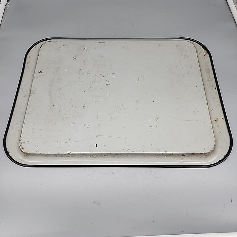 May include: A rectangular, off-white metal tray with a black border. The tray has a slightly raised edge and a worn appearance, suggesting it is vintage or has been well-used. The surface shows minor imperfections.