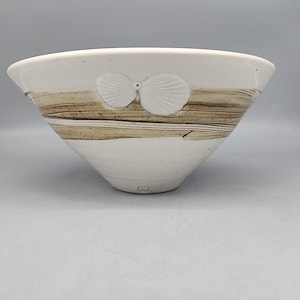 May include: A white ceramic bowl with a wide rim and a tapered base. The bowl features a brown, horizontal stripe design and a pair of white, shell-shaped embellishments. The bowl has a simple, elegant design.