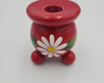 May include: A small, round, red wooden candle holder featuring a hand-painted white daisy with a yellow centre on the front. The object has a wide, flat top with a central circular opening and is supported by three small, spherical red feet.