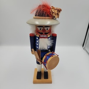 May include: A wooden nutcracker soldier figurine, featuring a white hat with a red feather and a pinecone. The soldier wears a blue jacket, white pants, and holds a drum. The base is made of wood.