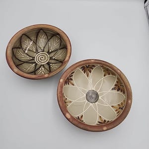 May include: Two handmade ceramic bowls with floral designs. The bowls have a brown rim and a beige interior. One bowl features a spiral design in the center, while the other has a flower design.