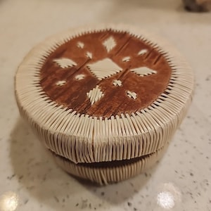 Peut inclure: Une boîte ronde faite à la main avec un couvercle. Le couvercle présente un centre brun veiné de bois avec des motifs géométriques blancs. Les côtés sont tissés avec un matériau de couleur claire, créant un effet décoratif texturé. La boîte est probablement utilisée pour le stockage.