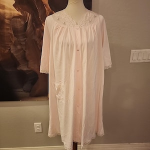 May include: A light pink, knee-length robe with short sleeves and a button-up front. The robe features a delicate lace trim along the neckline, sleeves, and hem. Small, decorative embellishments are placed on the neckline. A pocket is visible on the left side.