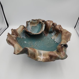 Op de afbeelding: Een keramische kom met een golvende, geplooide rand met een mix van bruine en blauwgroene glazuren. De binnenkant heeft een getextureerd, gespikkeld oppervlak, terwijl de buitenrand vloeiende bruintinten met wit en lichtblauw vertoont.