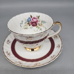 Op de afbeelding: Witte theekop en schotel set met een bloemenpatroon van roze en blauwe bloemen. De kop heeft een gouden oor en rand, met een bordeauxrode band. De schotel heeft een bordeauxrode ring en gouden accenten.