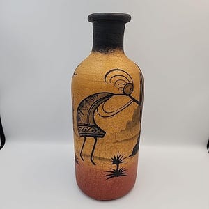 May include: A tall, decorative vase with a black rim and a textured, tan body. The vase features a black Kokopelli figure playing a flute, with desert landscape details. The base transitions from tan to a reddish-brown color.