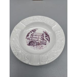 May include: White ceramic ashtray with a purple illustration of a water mill in the center. The ashtray has a decorative floral pattern around the rim and three notches for resting cigarettes. A vintage-style home decor item.