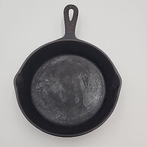 May include: A black cast iron frying pan with a long handle and a rounded cooking surface. The pan is round, with a diameter of approximately 20 cm. The handle has a hole for hanging.