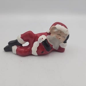 May include: A ceramic Santa Claus figurine in a reclining pose, dressed in a red suit with white trim and black boots. The figurine has a white beard and a smiling face, ideal for Christmas decorations.
