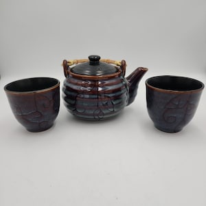 May include: A ceramic tea set consisting of a round teapot with a black lid and a bamboo handle, accompanied by two matching cups. The pieces feature a dark, glossy glaze with horizontal ridges and a subtle, swirling embossed leaf pattern.