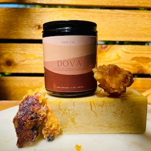 May include: A jar of "DOVA Miracle Healing Salve" with a black lid and a brown label. The label has the text "AKIV CO." and "100% ORGANIC | 240 ML". The jar is next to a block of yellow wax and pieces of amber-coloured resin.