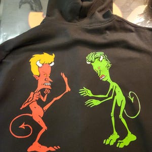 May include: Black hooded sweatshirt featuring a cartoon graphic of two devils. One devil is red with yellow hair, kicking. The other is green with green hair, reaching out. The sweatshirt has a drawstring hood.