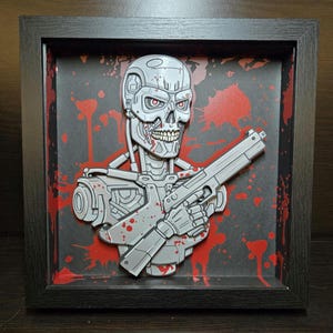 May include: A framed 3D papercraft of a Terminator character holding a gun. The character is made of gray paper and has red blood splatters on it. The background is black with red blood splatters.
