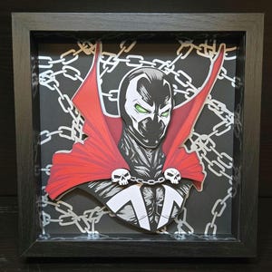 May include: A 3D papercraft of the comic book character Spawn. The character is depicted with a red cape, a black and white suit, and a skull on his chest. The background is a black and white pattern of chains.