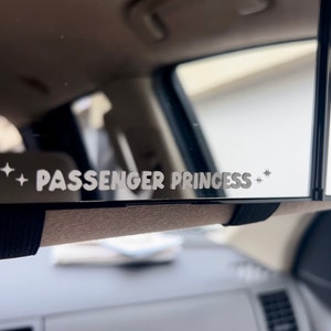 以下が含まれることがあります： 「PASSENGER PRINCESS」という文字と両側に星が2つある白いステッカーが貼られた黒いリアビューミラーです。