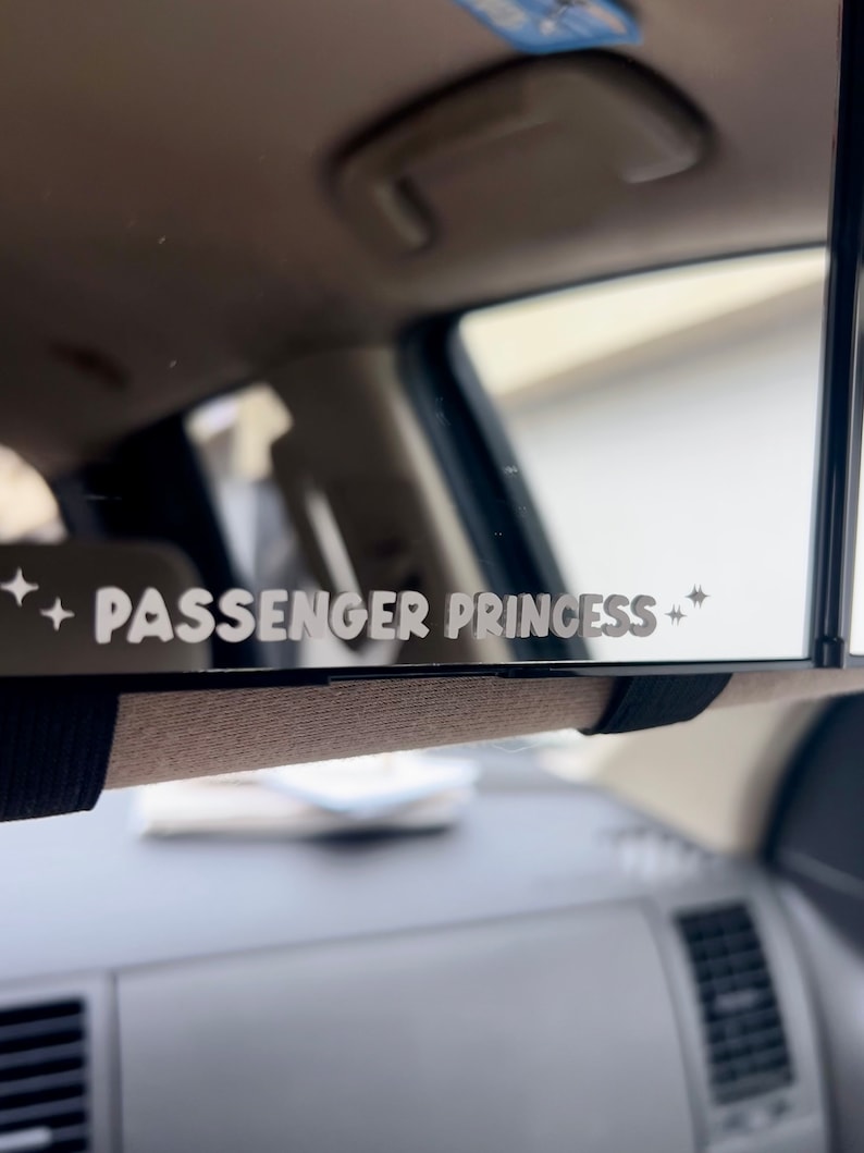 以下が含まれることがあります： 「PASSENGER PRINCESS」という文字と両側に星が2つある白いステッカーが貼られた黒いリアビューミラーです。