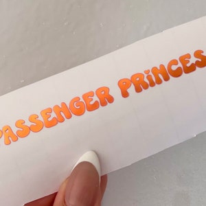 以下が含まれることがあります： オレンジ色のバブルレターで「...PASSENGER PRINCESS!」と書かれた白いステッカーです。