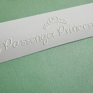 以下が含まれることがあります： 「Passenger Princess」というテキストが筆記体で書かれた白いステッカー。テキストの上と下に王冠と2つのダイヤモンドの形があります。