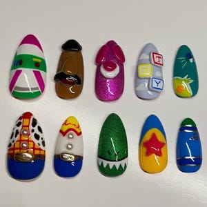 May include: A set of ten artificial nails, each uniquely designed with vibrant colors and cartoon-inspired themes. Designs include a train, a character with a mustache, a pink glittery character, and a blue nail with the letters T, Y, and Q.