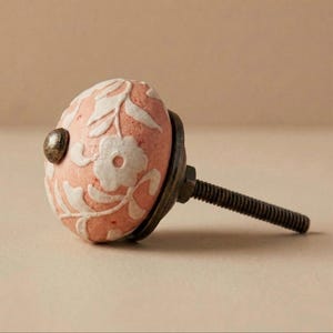 May include: A decorative cabinet knob with a pink ceramic finish and white floral design. The knob has a dark metal base, a small metal accent, and a threaded screw for installation.