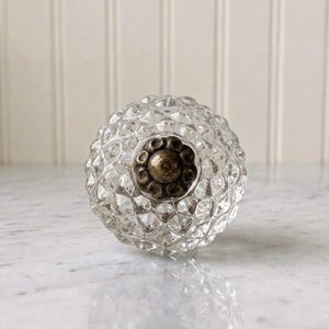 Op de afbeelding: Een ronde, heldere glazen kastknop met een textuur van een geometrisch diamantpatroon over het oppervlak. Het midden van de knop is geaccentueerd met een ronde, antiek afgewerkte bronzen metalen plaat met een geschulpte, bloemachtige rand.