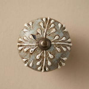 May include: A round, blue-grey ceramic cabinet knob with a white floral design. The knob has a metallic centre and is likely used for furniture. The design features white teardrop shapes radiating from the centre.