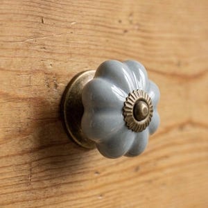 May include: A close-up of a light blue ceramic cabinet knob with a flower-like design. The knob has a brass base and center, mounted on a wooden surface with visible wood grain.
