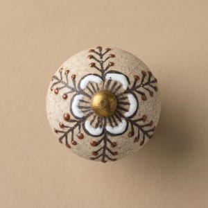 May include: A decorative cabinet knob with a beige, speckled finish. The knob features a floral design with white and brown accents, and a gold-colored center. The design is symmetrical and detailed, suitable for furniture or decor.
