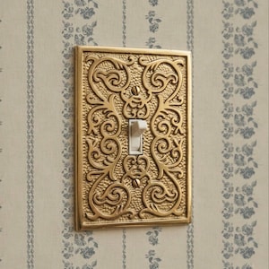May include: A decorative light switch cover with an ornate, gold-colored design. The cover features a rectangular shape with intricate scrollwork and a white toggle switch. The background is a floral wallpaper.