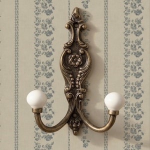 Puede incluir: Perchero de pared de estilo antiguo en tono bronce, con dos brazos curvos que terminan en pomos esféricos blancos. El gancho presenta un elaborado diseño floral y de volutas, ideal para un interior vintage.