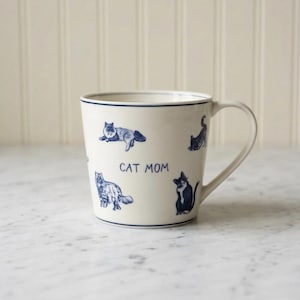 May include: A cream-colored ceramic mug featuring a series of blue illustrations of various cats in different poses. The text CAT MOM is printed in blue in the center. The mug has a thin blue rim and a single handle on the right side.