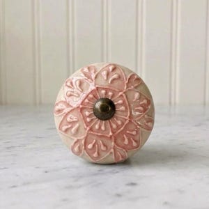 Może przedstawiać: Okrągła ceramiczna gałka meblowa z wypukłym, tłoczonym wzorem kwiatowym w kolorze delikatnego różu na kremowym tle. Środek gałki jest zaakcentowany małym, ciemnym, okrągłym metalowym elementem, tworzącym wyraźny kontrast.