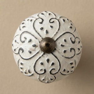 May include: A white, round cabinet knob with a distressed finish. The knob features a black, ornate floral design and a bronze-colored center. This decorative hardware adds a vintage touch to furniture.