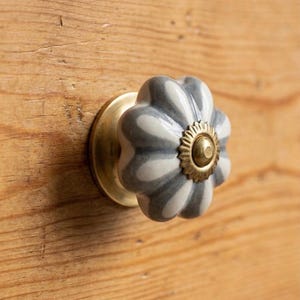 May include: A close-up of a decorative cabinet knob. The knob has a flower-like shape with gray and white stripes. It is mounted on a brass base and set against a wooden surface.