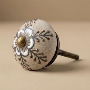 Könnte beinhalten: Dekorativer Keramik-Schrankknopf mit floralem Design. Der eiförmige Knopf hat einen beigefarbenen Hintergrund und ein weißes und braunes Blumenmuster. Er hat eine messingfarbene Mitte und eine Gewindeschraube aus Metall zur Montage.