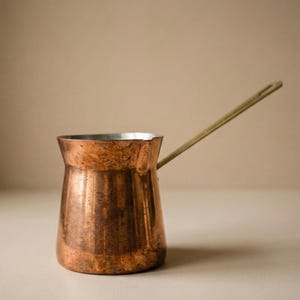May include: A copper coffee pot with a long, thin handle. The pot has a wide base that tapers to a narrower neck and a flared rim. The copper has a warm, reddish-brown color with a slightly textured surface. The handle is a lighter, golden color.