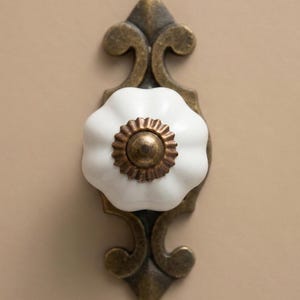 May include: A decorative cabinet knob featuring a white, flower-shaped ceramic handle and a bronze-coloured metal base. The base has an ornate, vintage design. The knob is mounted on a neutral-toned surface.