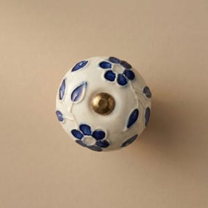 May include: A white ceramic cabinet knob with a floral design. The knob features blue flowers and leaves, with a brass-colored center. The design is raised, giving it a three-dimensional appearance. The knob is a decorative element for furniture.