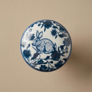 Puede incluir: Pomo de armario redondo de cerámica blanca con un diseño floral y de conejo azul. El conejo está en el centro, rodeado de flores y follaje azules. El pomo tiene un borde azul.